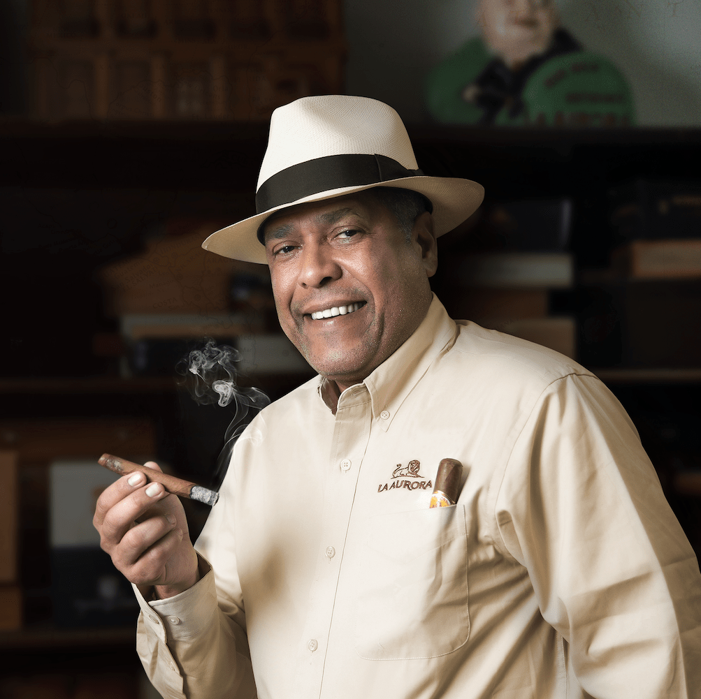 Man in beige shirt and white hat holding a lit cigar, smiling with a blurred background.