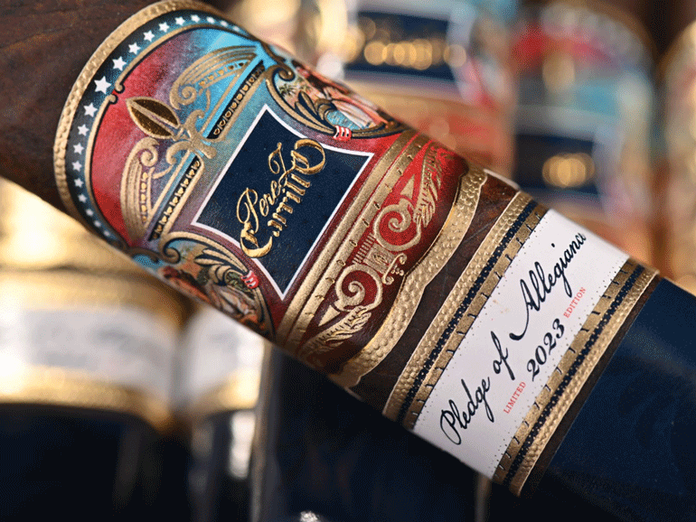 Close-up of a decorative cigar band featuring intricate designs and text reading "Perez Carrillo Pledge of Allegiance Limited 2023 Edition" on a dark background.